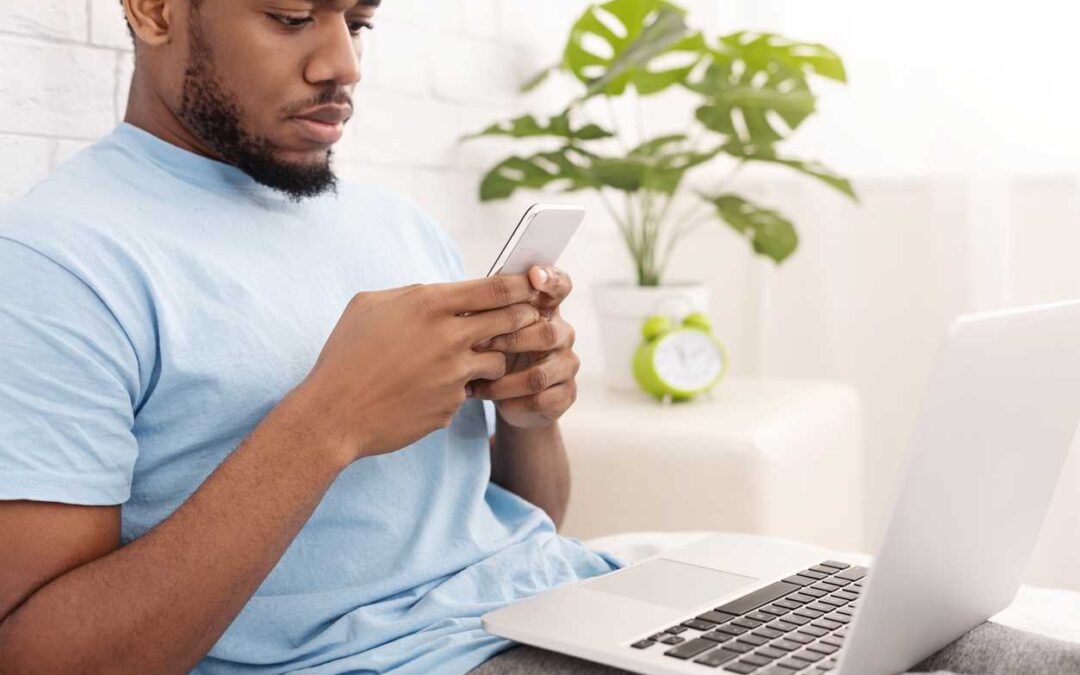 A man relaxing on a bed, typing on a smartphone, and holding a laptop.