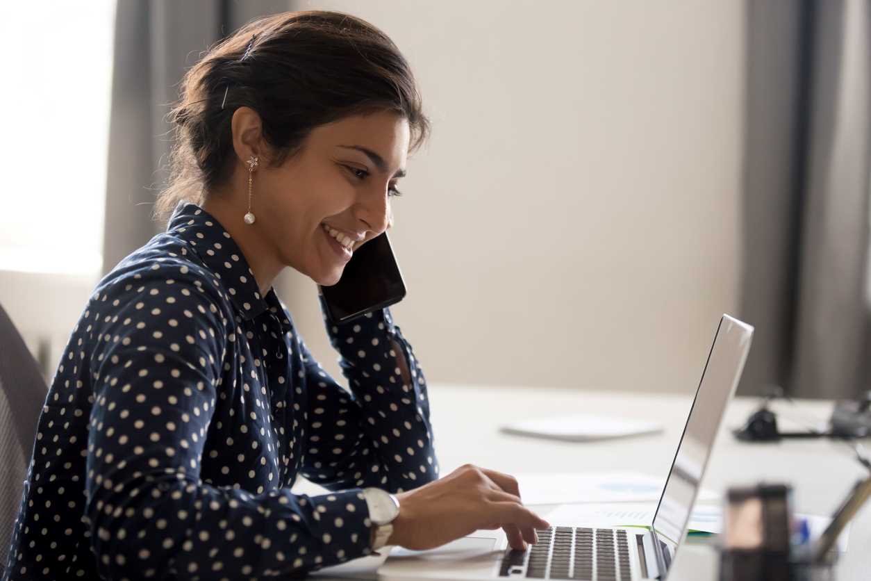 Smiling business woman talking on a mobile phone while typing on a laptop.