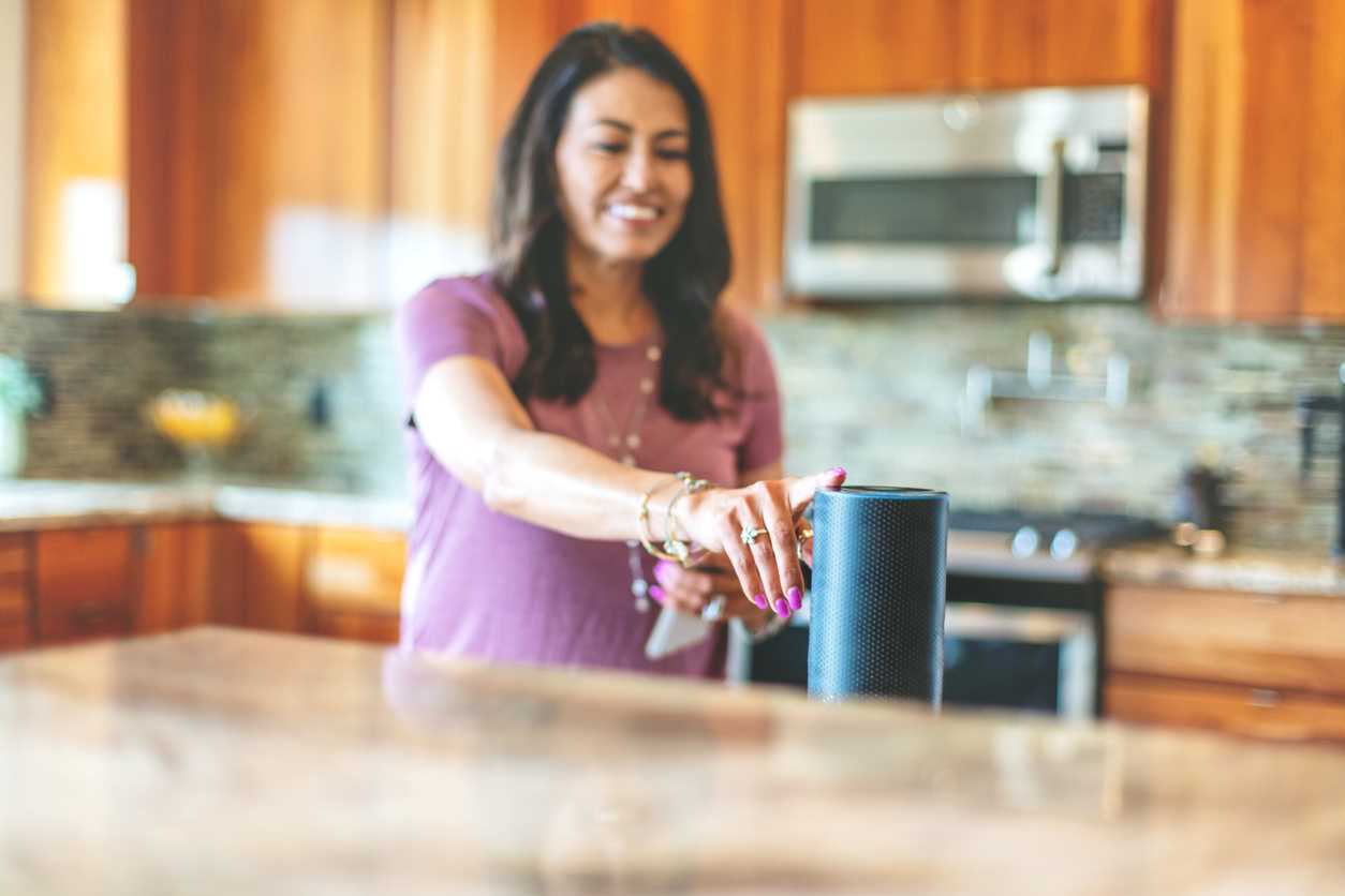 A woman standing in a home kitchen touches a smart speaker and practicing security for smart home.