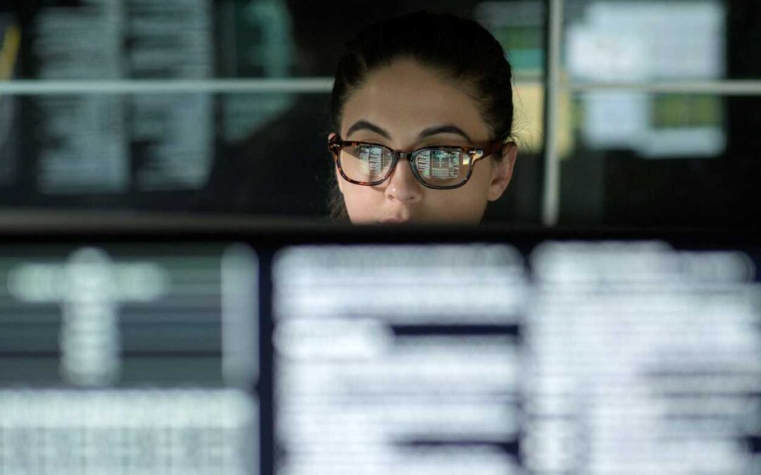 Woman wearing eyeglass sitting down with blurred computer screens all around her.