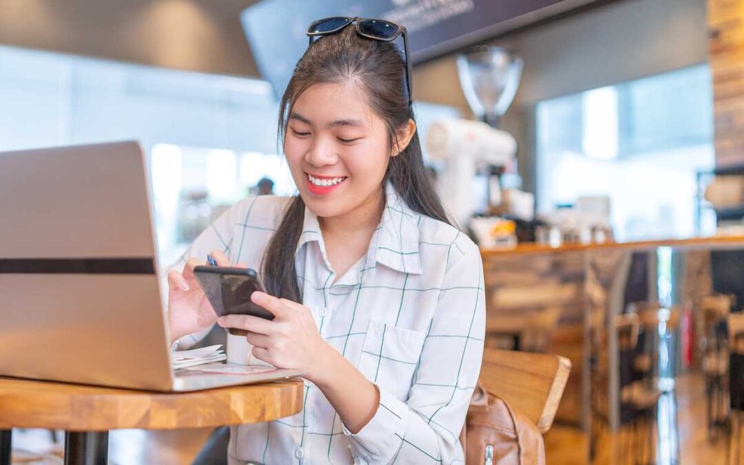 A woman working in a coffee shop. She smiles as she navigates her smartphone.