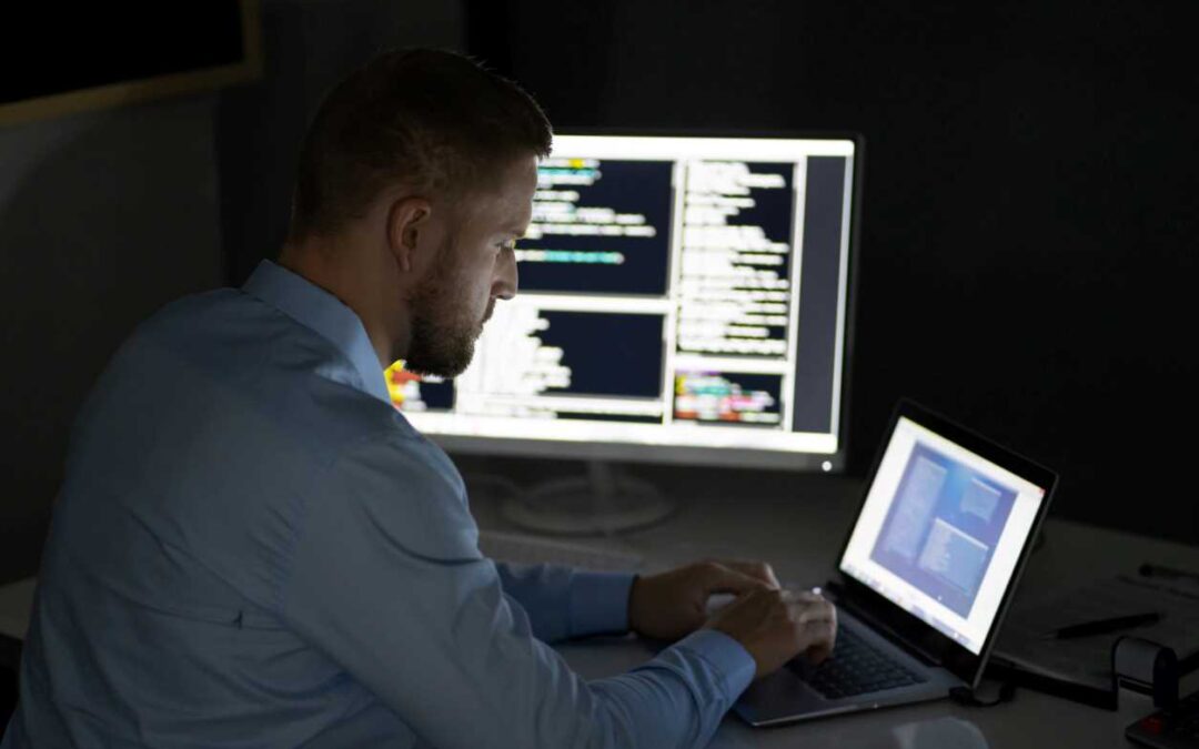 Man in a dress shirt typing on a laptop next to a larger computer monitor displaying data.