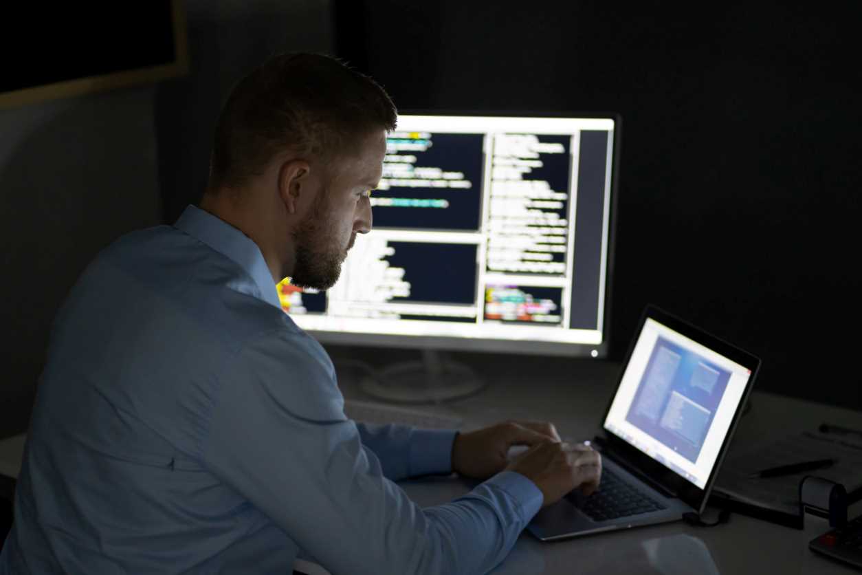 Man in a dress shirt typing on a laptop next to a larger computer monitor displaying data.