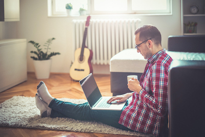 Young man sitting on the floor drinking coffee and surfing the internet using a laptop.