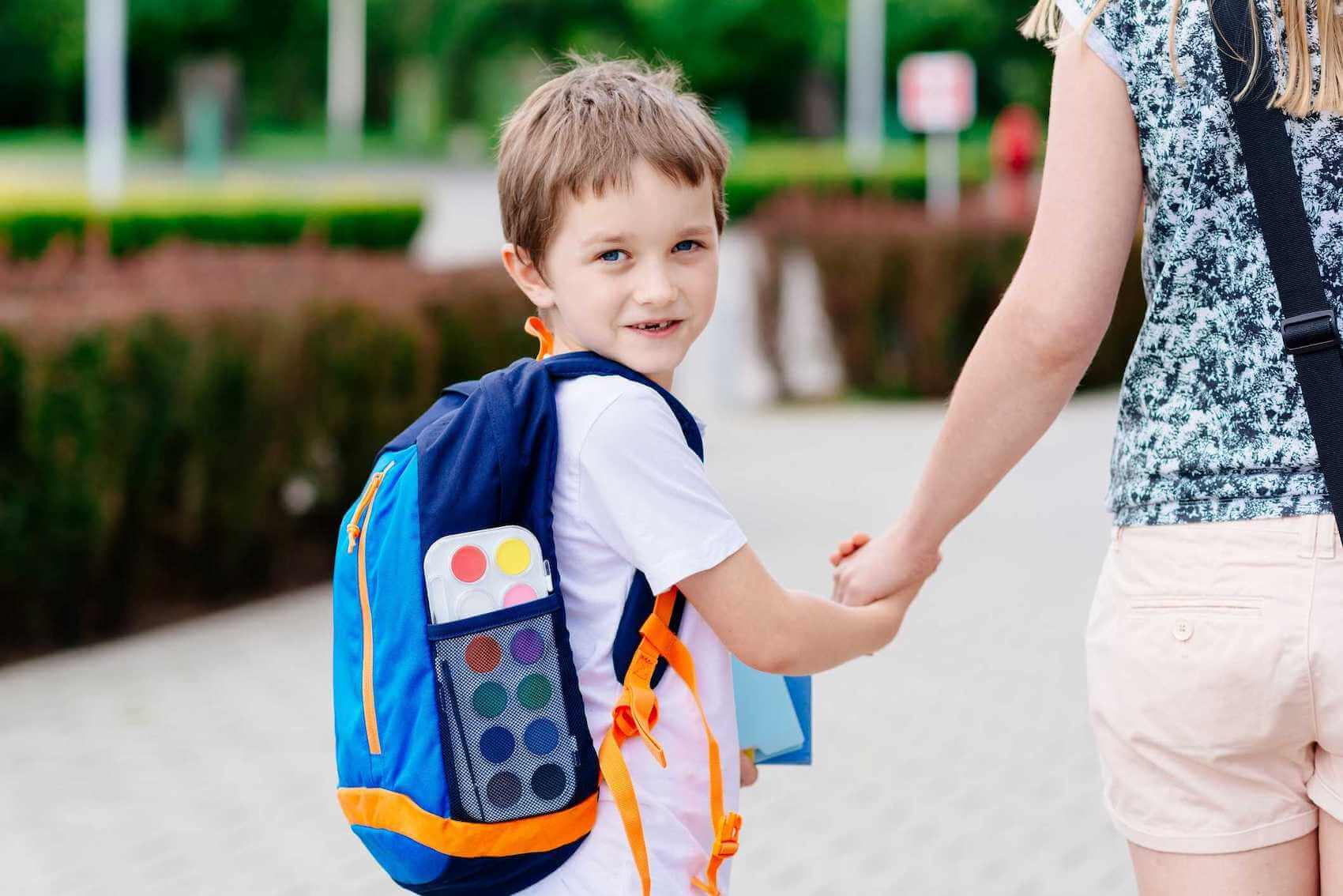 Young boy walking & wearing a backpack as a woman holds his hand.