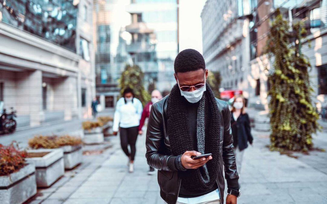 A man wearing a face mask walks down a city sidewalk as he looks at his smartphone.