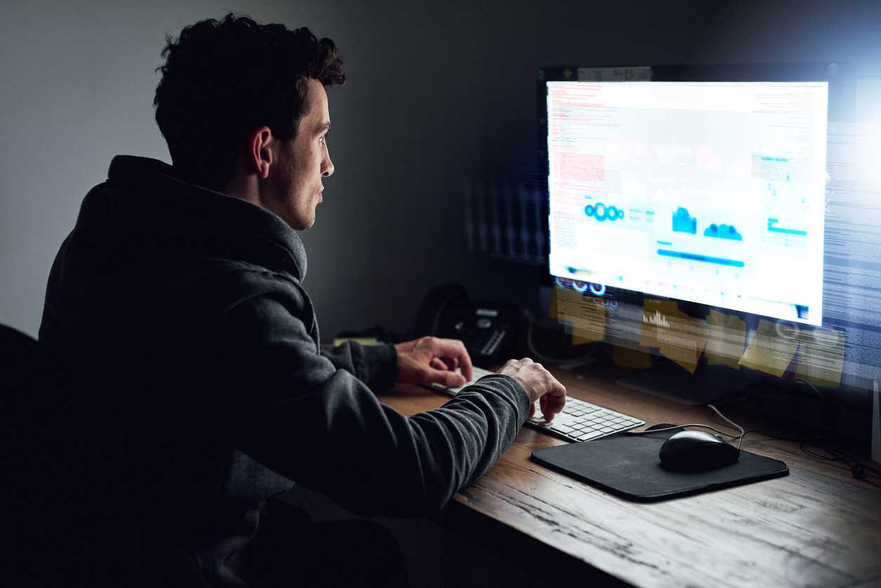 A hacker sitting at a desk and viewing data on a large computer screen.