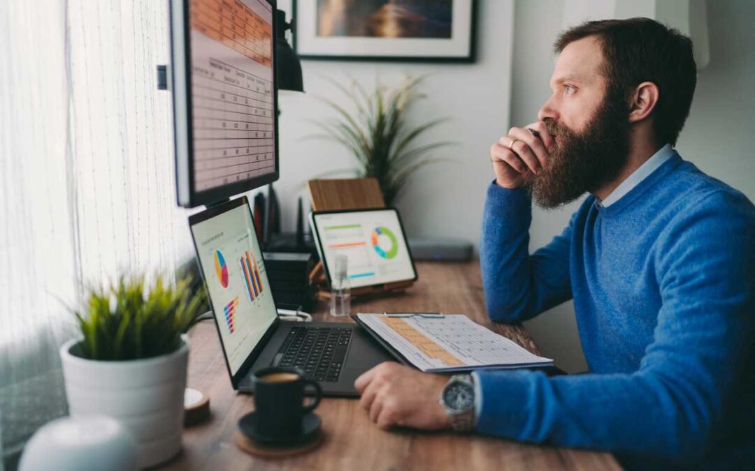 A man is looking at spreadsheets and graphs on 3 computer monitors as he sits at a large office desk.