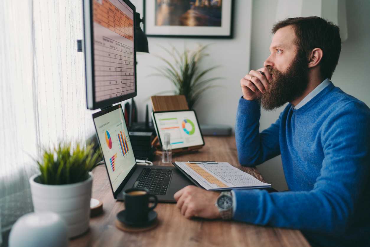 A man is looking at spreadsheets and graphs on 3 computer monitors as he sits at a large office desk.