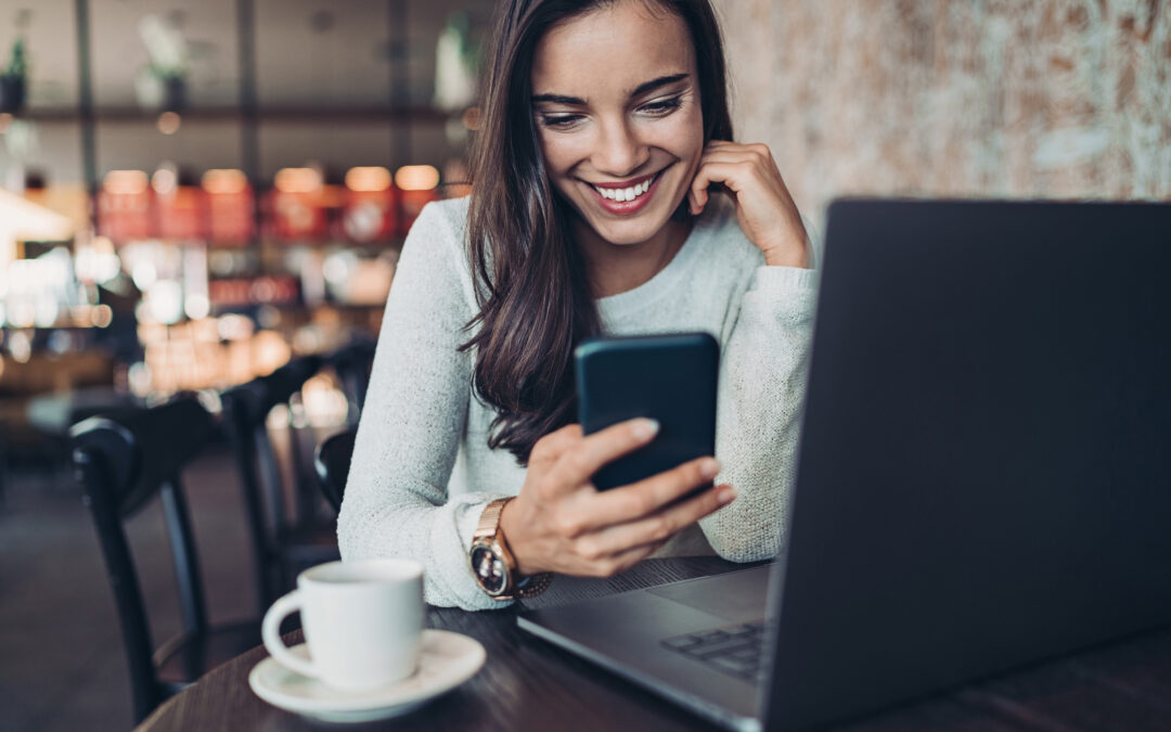 Smiling woman looking at cell phone in cafe. She has a laptop open and a cup of coffee nearby.