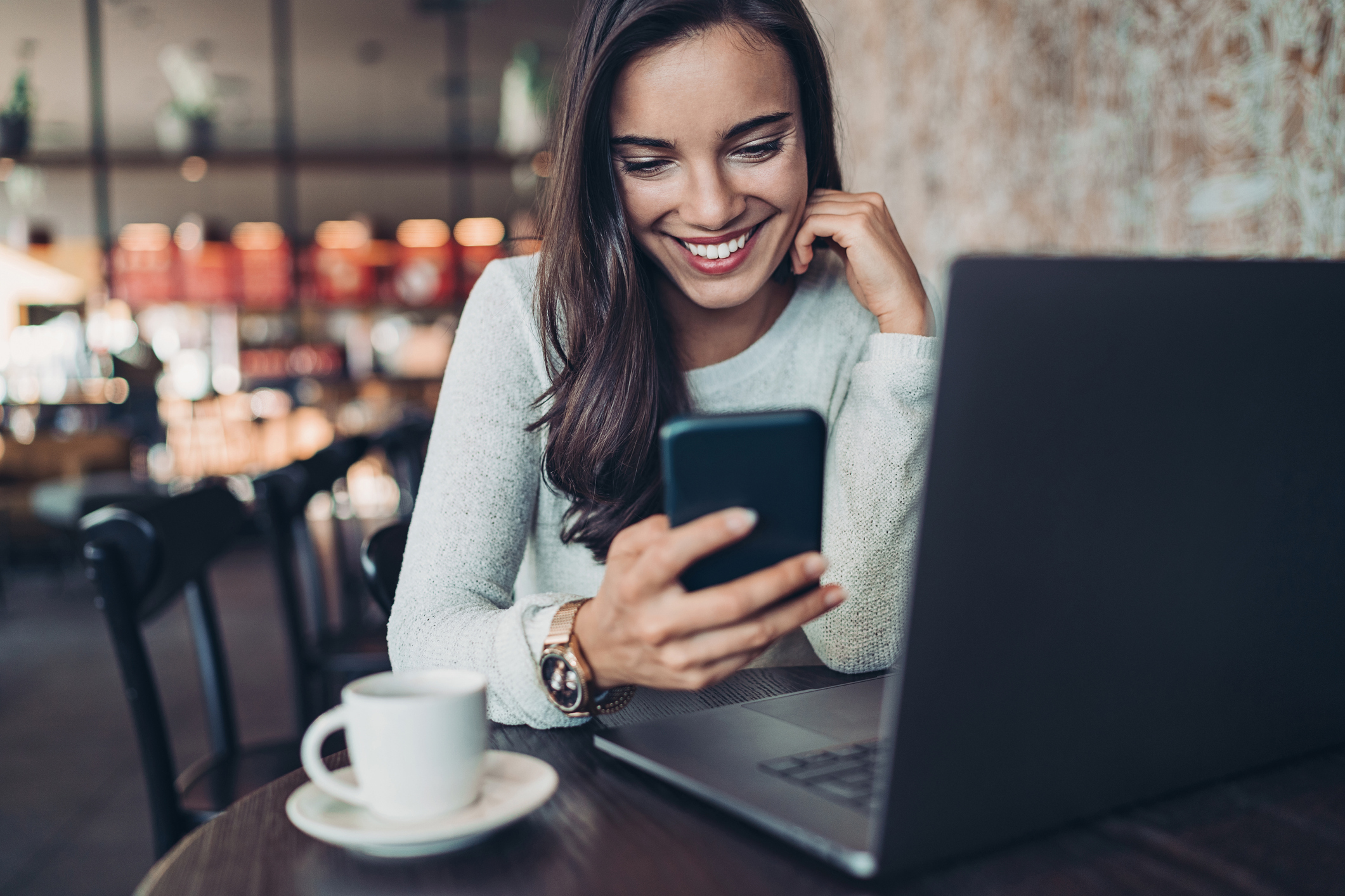 Smiling woman looking at cell phone in cafe. She has a laptop open and a cup of coffee nearby.