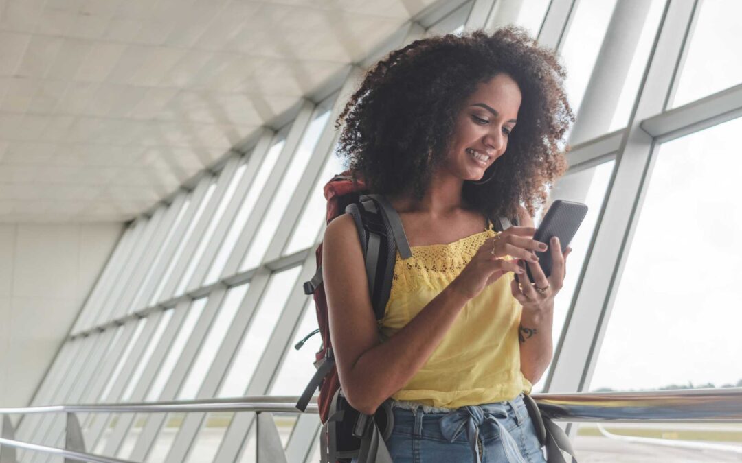 Traveling woman in an airport wearing a backpack and looking at her cell phone to avoid a travel scam.