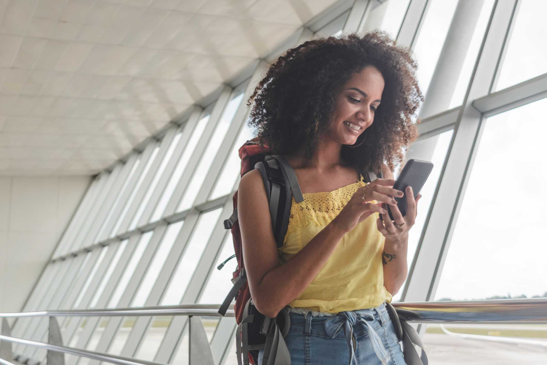 Traveling woman in an airport wearing a backpack and looking at her cell phone to avoid a travel scam.