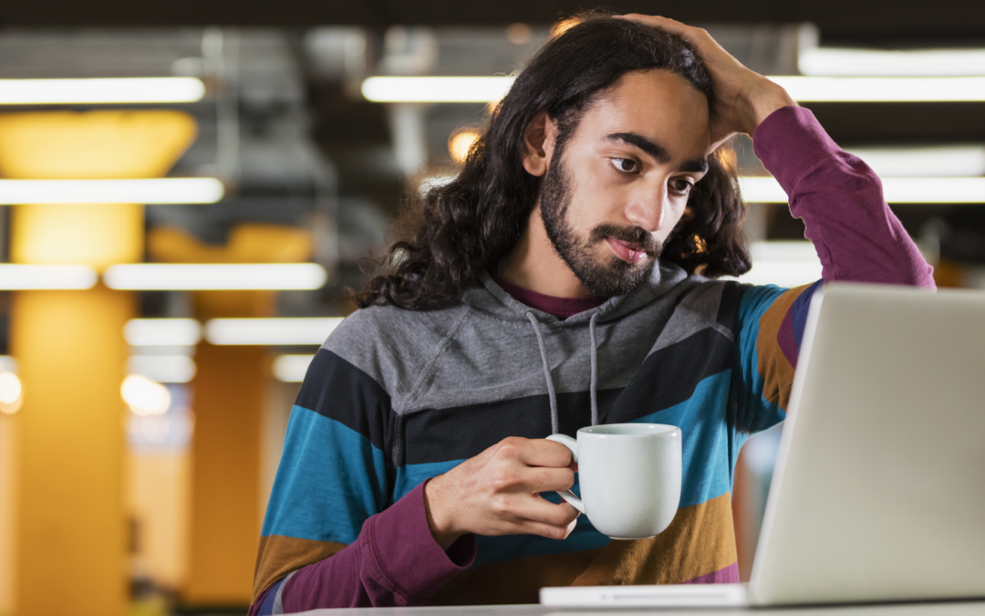 Man with long black hair is holding a coffee cup and looking at a laptop screen.