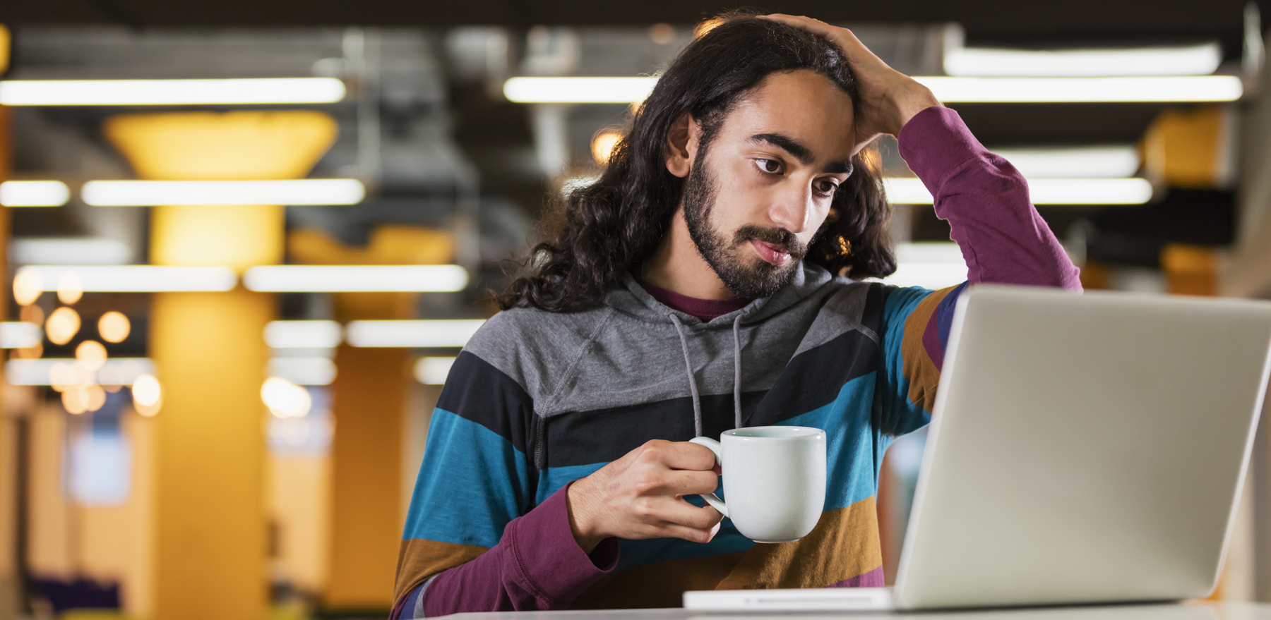 Man with long black hair is holding a coffee cup and looking at a laptop screen.