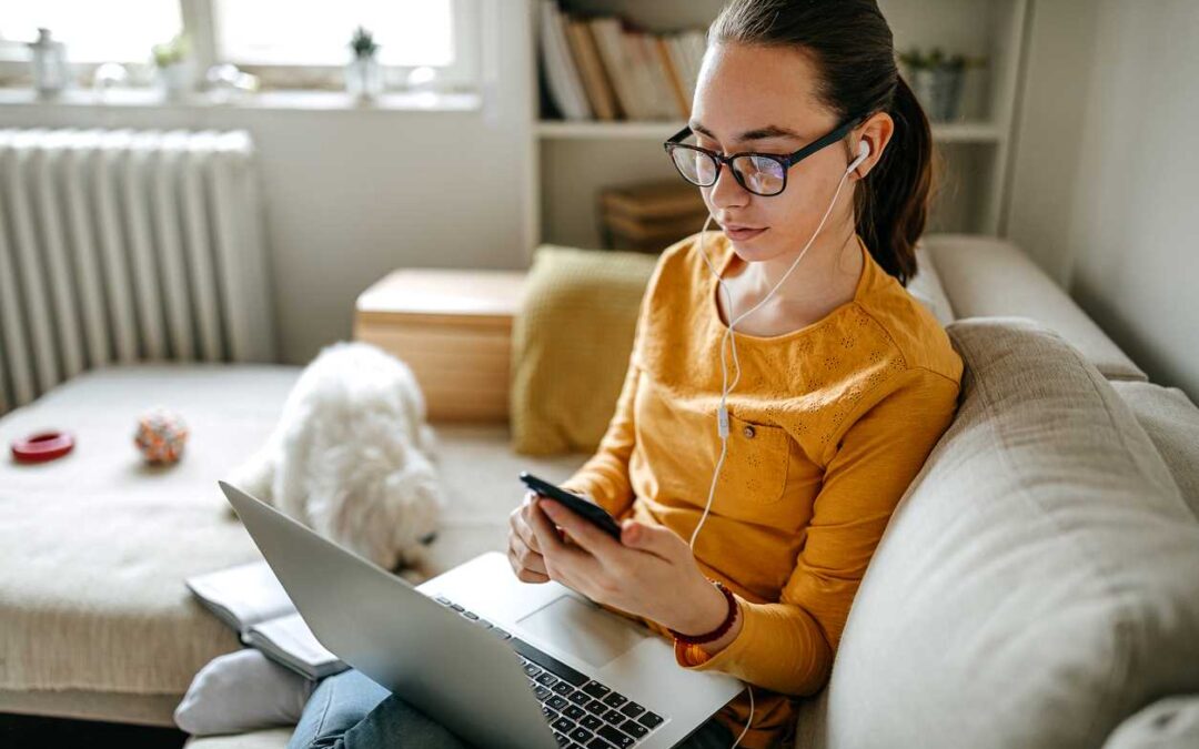 Woman casually sitting on her sofa, looking at mobile phone and holding a laptop. She is wearing earphones and a small white dog plays nearby on the sofa.