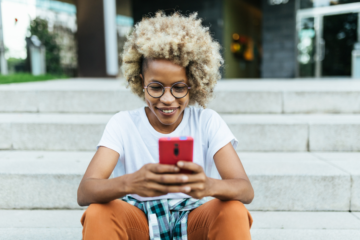 Smiling woman viewing Facebook on her mobile phone while sitting on building steps outside.