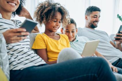 Young girl viewing a computer tablet with her brother as she sits on a sofa near her mother and father.