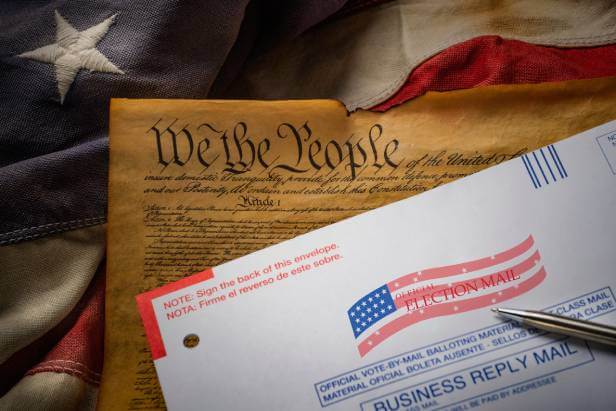 An official U.S. vote by mail ballot envelope next to a flag, ink pen and a copy of the U.S. Constitution.